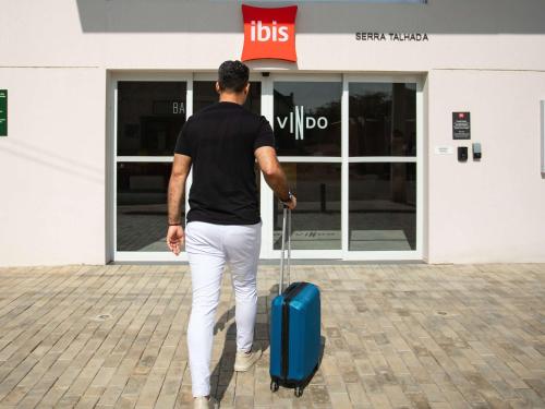 a man pulling a blue suitcase in front of a store at Ibis Serra Talhada in Serra Talhada