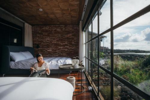 a woman sitting in a room with a bed and a window at Ocean View Riad Jeju in Jeju