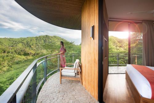 a woman standing on a balcony with a bed and a chair at Suitree Experience Hotel in Guanacaste