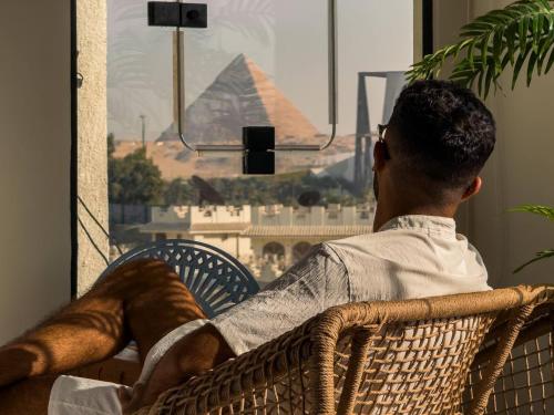 a man sitting in a chair looking out a window at the pyramid at Fancy Pyramids View Khan Giza's Gem in Cairo