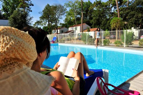 2 personnes assises sur une chaise pour lire un livre au bord de la piscine dans l'établissement Résidence Goélia Domaine des Oyats, à Longeville-sur-Mer