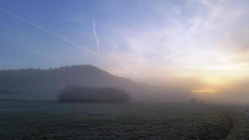 a foggy field with the sun rising behind a hill at Luisenhof in Geisfeld