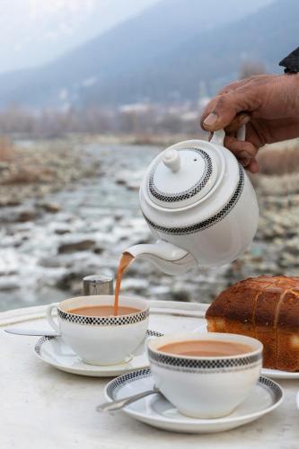a person pouring coffee into two cups on a table at Water's Edge by VEO - Part of StayVista in Chak-i-Dāra