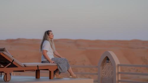 a woman sitting on a bench in the desert at Stars Gate Camp in Al Wāşil