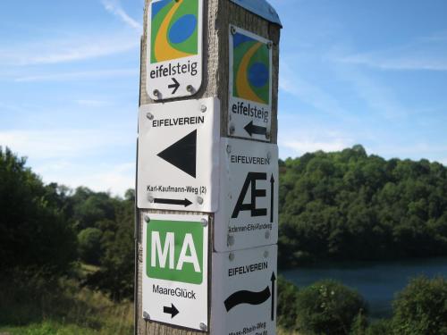 a pole with signs on it with a lake in the background at Ferienwohnungen Haus Hinneres in Schalkenmehren