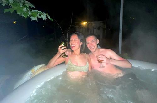 a man and woman sitting in a bath tub with wine glasses at The Stabbin Cabin on Grant Island, Brantingham Lake - HotTub, Boat, Pet Friendly in Glenfield