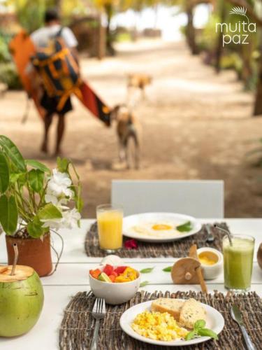 een tafel met borden eten en een hond op de achtergrond bij Muita Paz Rooms in Atins