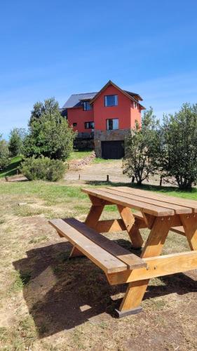 a wooden picnic table in a field with a red house at Complejo Don Cirilo in Villa Pehuenia