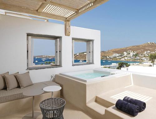 a soaking tub in a room with a couch and windows at Kove Mykonos - A Myconian Collection Hotel in Ornos