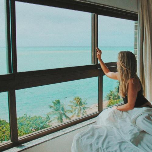 a woman sitting on a bed looking out a window at the beach at Hotel Ponta Verde Maceió in Maceió