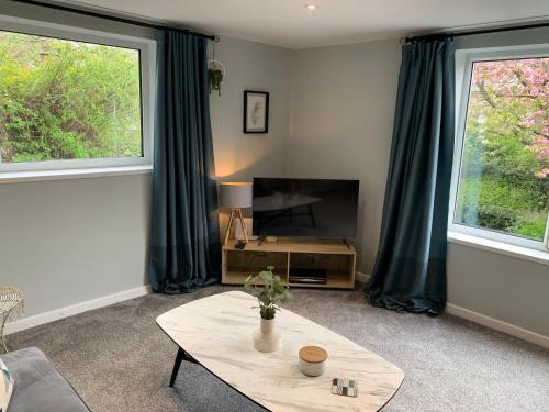 a living room with a table and a tv and two windows at Bronte Country - 2 Bed Apartment near Cliffe Castle in Keighley