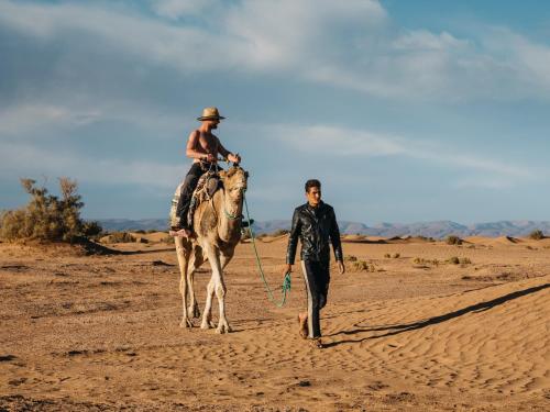 a man walking a camel with a man riding it in the desert at Bivouac Les Nomades & Foum zguid to chegaga tours in Foum Zguid