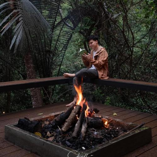 a man sitting on a bench next to a fire at บ้านเคียงธาร โฮมสเตย์ แม่แมะ Cascade Lodge in Amphoe Chiang Dao