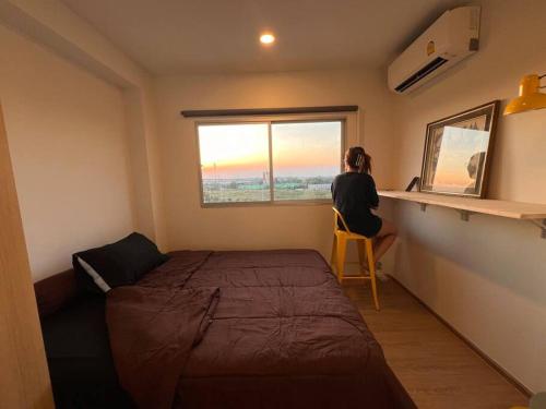 a woman sitting in a chair in a bedroom looking out the window at Triple Cats Condo in Samutprakarn
