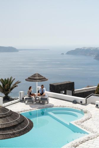 two people sitting under an umbrella next to a swimming pool at Santorini Princess Spa Hotel in Imerovigli