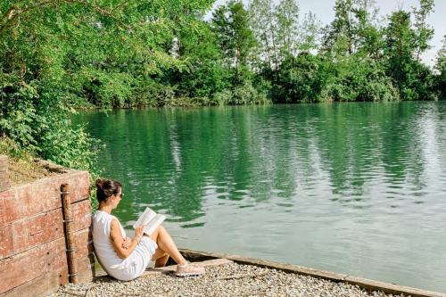 a woman sitting next to a lake reading a book at Amy B&B casetta di charme in Vercelli
