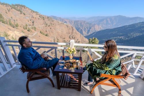 a man and woman sitting at a table on a balcony at SaffronStays Alpine blue-6 bhk villa with private deck and valley views from each room in Shimla