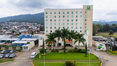 a large white building with palm trees in front of a city at Holiday Inn Uruapan by IHG in Uruapan del Progreso