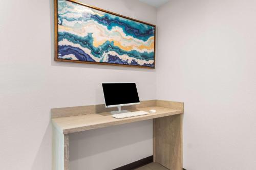 a desk with a computer on a wall with a painting at Clarion Pointe - Fort Worth Arlington West in Fort Worth