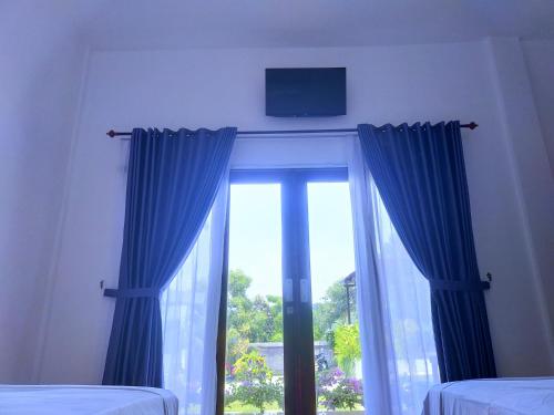 a bedroom with a window with blue curtains and a tv at The Local Are Guling in Kuta Lombok