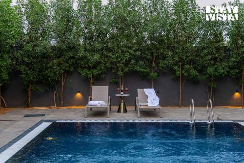 a pool with two chairs and a table and trees at StayVista at Urban Escape - 2 Pools, Lawn & WiFi in Jaipur