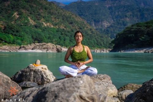 a woman is sitting on rocks in front of a body of water at Zostel Rishikesh, Tapovan in Rishīkesh