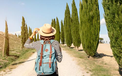 a woman with a backpack walking down a dirt road with trees at Abbadia Sicille Relais in Trequanda