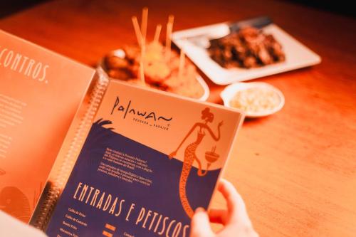a person holding up a book in front of a table with food at Palawan Pousada & Bistrô in Nísia Floresta