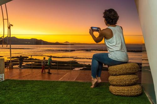 a woman sitting on a stack taking a picture of the sunset at OceanSound White in Las Palmas de Gran Canaria