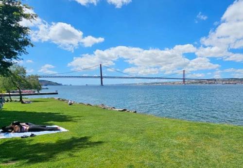 a person laying on the grass near the water with a bridge at Big apt 4beds with terrace, 15 mins to Lisbon in Almada