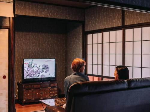two people sitting in a living room watching a television at ひかりの森Inn in Kumamoto