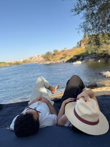 two people laying on a blanket next to a body of water at Nala Narty Boutique Hotel & Farm in Aswan