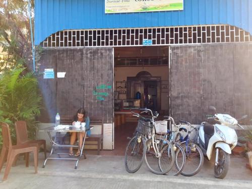 a woman sitting at a table outside of a building with bikes at SunRise Villa in Kampong Cham