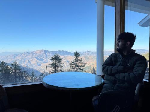 a man sitting next to a table in front of a window at The Sky Mountain Kufri I Serviced Villa I Balcony I Valley and Mountain View I Family Groups & Bonfire I Memories & Enjoyment Villa I Operated Charvi Retreat by Exotic Stays in Kūfrī