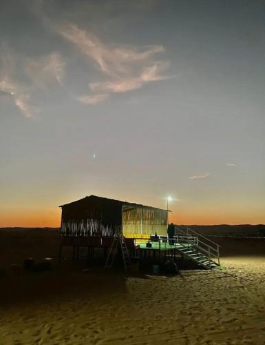 a shack on a beach with the sun setting at مخيم رملة الجوفاء Al-Jawfa Dunes Camp in Al Wāşil