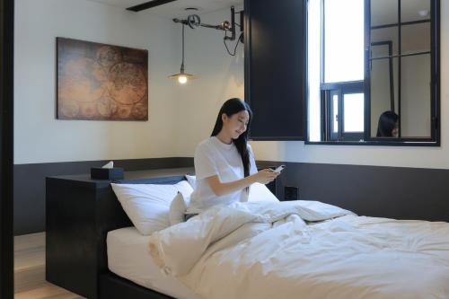 a woman sitting on a bed looking at her cell phone at Hotel Yaja Siheung Jeongwang in Siheung