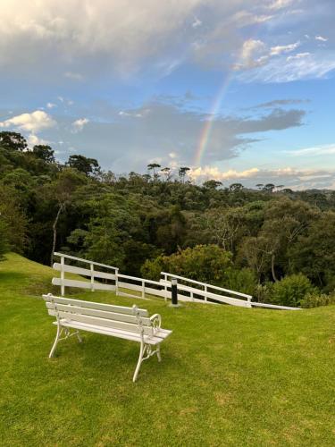 a white bench sitting in the grass with a rainbow in the background at Ape 2 Qts - Campos do Jordão - Condomínio Alto da Boa Vista - Varanda com Vista Panorâmica - Apartamento Inteiro in Campos do Jordão