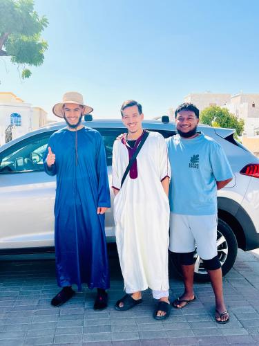 three men are standing in front of a car at Sukoon Hostel in Muscat