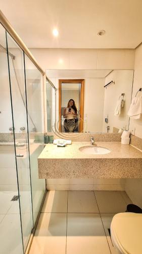 a woman taking a picture of a bathroom with a sink at Coruja Imob - Flat Crystal Place in Goiânia