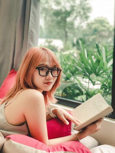 a woman with glasses reading a book in front of a window at Kayla Homestay - Bungalow Rose Double in Ba Vì