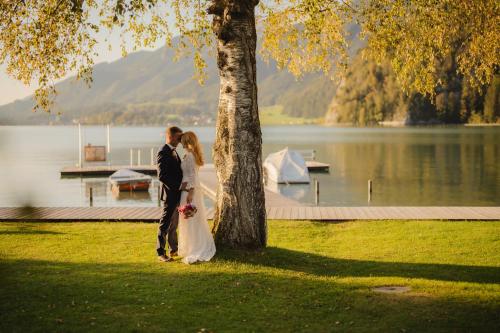 a bride and groom standing next to a tree next to a lake at Seehotel Brandauer's Villen in Strobl