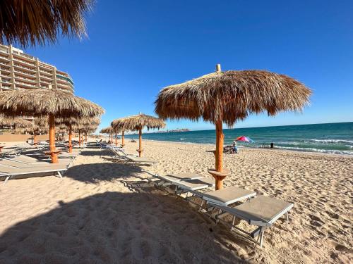 a beach with many straw umbrellas and chairs and the ocean at Paradise Found Luxe 2BR Condo in Iconic Location in Puerto Peñasco