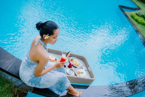 a woman sitting in front of a pool with a drink at Abhirama Villas and Spa in Ubud