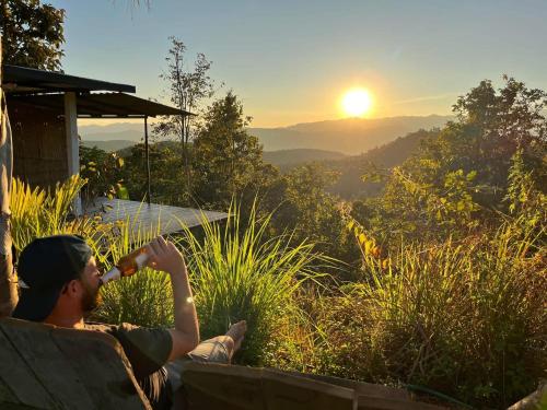 a man is sitting on a bench drinking a drink at Monsonkeang Camping view ม่อนสันเกี๋ยง in Mae Chaem