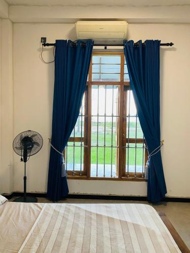 a bedroom with a window with blue curtains and a fan at Lago Inn in Tissamaharama
