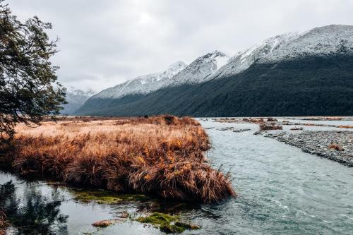 a river with grass and snowy mountains in the background at The Bealey Hotel in Arthur's Pass