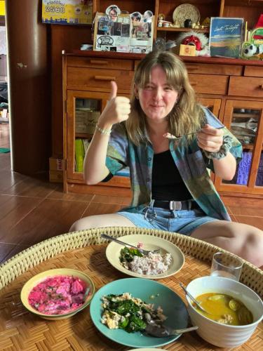 a woman giving a thumbs up while sitting at a table with food at โฮมสเตย์บ้านนายสิงโตเขาคูหา in Ban Khu Ha Nai (1)