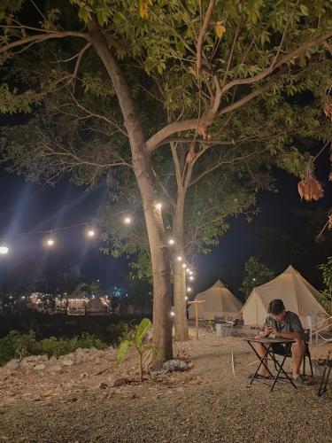 a man sitting at a table under a tree at night at Tháng Tư Glamping in Ha Tien