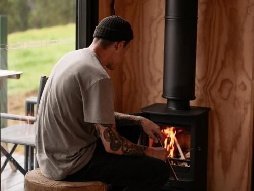 a man with a tattoo on his arm sitting in front of a stove at Off-Grid Cabin with Valley Views and Hot Tub - Velma in Tarra Valley