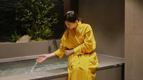 a woman in a yellow robe drinking water from a fountain at 스테이 패스포트 후쿠오카 료칸 대전 유성점 in Daejeon